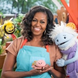 Michelle Obama smiling with the TV-show characters, Waffles and Mochi, beside her.